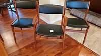 Three teak frame chairs with black upholstered seats and backs, two armless side chairs and one armchair on hardwood floor near couch and rolled rug.