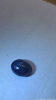 Close-up photo of the oval natural blue star sapphire gemstone lying on a flat surface showing its dark blue color and cabochon dome shape.