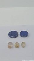 Five polished oval natural gemstones on white background, two Lapis Lazuli deep blue stones on top row, three golden rutilated Quartz stones below.
