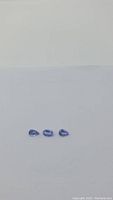 Three blueish purple tanzanite gemstones laid in a row on white background, visible oval and pear shapes.