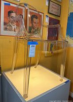 Front and side view of transparent Louis Ghost Chair on a pedestal against a wall with portraits and posters.