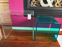 Clear and green-tinted Lucite side tables placed side by side on a wooden floor against a pink wall.