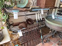 Front view showing the full three-tier wrought iron shelf with various outdoor decorative items around it, highlighting the rusty spots and dimensions.