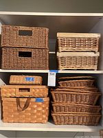 Photo of mixed baskets on shelving showing various sizes and colors including dark brown rectangular woven baskets with cut out handles, medium brown wicker baskets with wooden edges, multiple large light brown woven baskets with handles, and several woven trays.