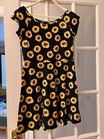 Front and back views of a black dress with a yellow sunflower print, short sleeves, fitted waist, and flared skirt on a wooden hanger, hanging on a door.