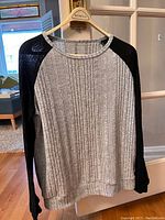 Front view of the medium soft grey and black sweater hanging on a hanger, showing ribbed grey body with black sleeves.