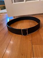 Black belt arranged in a circle with a silver rectangular metal buckle on a wooden floor.