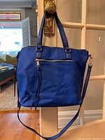 Front view of a royal blue bag with two handles, a front zippered pocket, and an adjustable shoulder strap hung on a door handle.