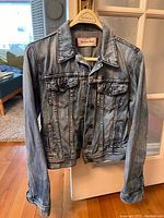 Front view of light blue Sunday Best women's denim jean jacket on hanger, showing button closure and two chest flap pockets with buttons.