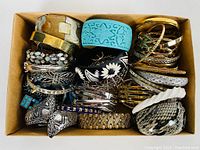 Top view of a cardboard box filled with a variety of estate bangles and bracelets showing colors, textures, and materials.