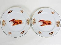 Top-down view of the pair of white bone china plates each decorated with a central lobster and surrounding seashells, gold trimmed edges visible.