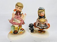 Two Japanese Hummel-style ceramic figurines shown side by side, depicting a girl with a chicken and another girl with a pie and dog.