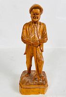 Frontal photo of handcrafted wooden sculpture of an old man holding a garden fork, standing on a wooden base.