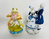 Front view of both figurines: dancing couple in blue and white, frog in yellow jacket on lily pad