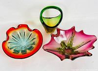Three Murano glass pieces: a red, orange and teal bowl with a sunburst pattern, a pink and green flower-shaped bowl, and a green and yellow vase with a loop handle.