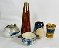 Five handcrafted pottery pieces arranged in one row including a small rounded pot, tall slender vase, beige vase with blue glaze, multicolored cup, and blue floral handled bowl.