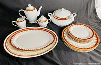 View of full vintage Royal Vienna porcelain set including coffee pot, sugar bowl, creamer, covered serving bowl, large platter, and stack of plates with decorative red and gold edges.