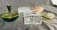 Five vintage trinket boxes displayed on gray fabric background, showing top and side views of each box including green glass, clear glass, heart-shaped, cat face, and gold-toned