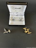 Three pairs of cuff links: silver-tone in Colibri case, gold-tone rectangular patterned pair, plain silver-tone round pair on black surface.