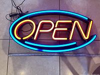 Photo of the neon OPEN sign laid flat on a tiled floor, showing bright orange neon letters spelling 'OPEN' inside a blue oval neon border.