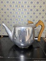 Side view of the teapot showing the polished metal body, wooden handle, and wooden lid knob against patterned wallpaper.