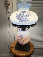 Photo showing a rare antique table lamp with white glass and blue floral designs, a two-switch mechanism, ornate dark metal base, and frosted glass top shade with wavy edges. Lamp is powered on with visible power cord.