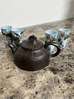 Brown textured Asian style teapot with sculpted lid knob and handle, set on a marble surface with four painted porcelain cups at the back.