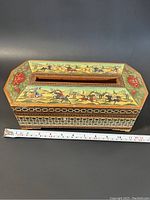 View of the top of the antique tissue holder showing horse rider scenes and floral painted design with measuring tape showing length