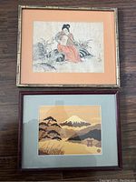 Photo showing two framed Asian paintings side by side on a wooden floor. One painting is of a seated woman holding a fan, the other is a landscape scene with Mount Fuji.