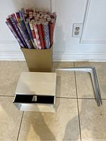 Full view of assorted rolled gift wrapping papers in a box, white metal storage rack, and metal toilet paper hanger.