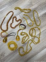 Photo showing 11 assorted necklaces laid out on wooden floor, various amber and yellow tones with different bead shapes and sizes.