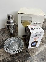 Close-up of stainless steel cocktail shaker, boxed wine glass, metal ashtray with embossed pattern, and boxed LED clock.
