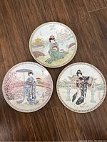 Three Japanese export plates arranged on floor showing different Geisha scenes with floral and architectural features.