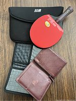 Photo of red rubber table tennis paddle, black padded case with Chinese logo, and two brown textured leather passport holders