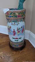 Full view of the oriental porcelain umbrella stand showing multicolor traditional figure painting and floral patterns on a wooden base.