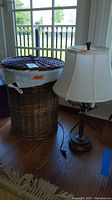 Photo showing wicker hamper with cotton insert next to metal base table lamp with shade near window on wood floor.