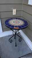 Round outdoor table with colorful mosaic tile top and black metal base positioned in a corner.