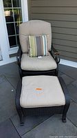 Front view of wicker lounge chair with beige cushions and matching ottoman on patio surface