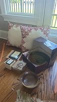Photo showing two throw pillows with red floral embroidery, two signed pottery bowls including one in original box, Willow Tree plaque, and two picture frames on wooden floor near window.