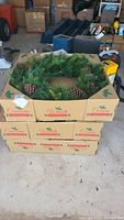 Stacked cardboard boxes containing three artificial pine and pinecone Christmas wreaths, shown on garage floor with surrounding tools and equipment.