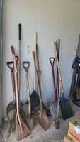 The entire collection of yard tools grouped and leaning against a white wall. Visible are multiple shovels, rakes, brooms, and a sledgehammer.