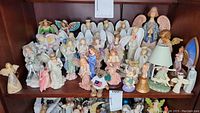 Wide view of multiple angel figurines arranged on two shelves inside a cabinet showing varying shapes and sizes.