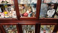 Wide view of several angel figurines inside a wooden glass cabinet, showing a variety of colors, sizes, and styles including classical angels and cherubs.