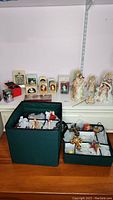 Wide view showing Christmas ornaments and figurines including angels, crosses and Lenox Holy Family displayed on a table with two green fabric storage bins containing decorations wrapped in paper and bubble wrap.