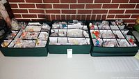 Three green compartmentalized storage bins filled with assorted Christmas ornaments, many individually wrapped in bubble wrap, showing a variety of characters and themes including Wizard of Oz, Snoopy, Disney, and more.