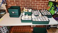 Three green fabric storage boxes on a table against a brick wall, two open compartment trays filled with wrapped ornaments and one empty large bin.