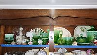 Wide shot of wooden shelf displaying green glassware, milk glass, china plates and small collectibles