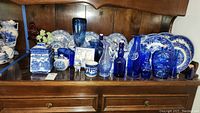 Wide view showing multiple Liberty Blue porcelain plates, blue glass vases, bottles, jars, and serving pieces arranged on wooden shelf.