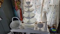 Wide view of collection of milk glass items including three-tiered serving stand, handled basket, and assorted vases and compote dishes arranged on a surface near window.