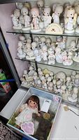 Multiple shelves filled with white and pastel-colored Precious Moments ceramic figurines with teardrop eyes, depicting children and angels in various poses, plus a boxed Precious Moments Laura doll at the bottom.